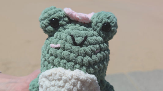 Green crochet frog toy with pink accents held against a neutral background