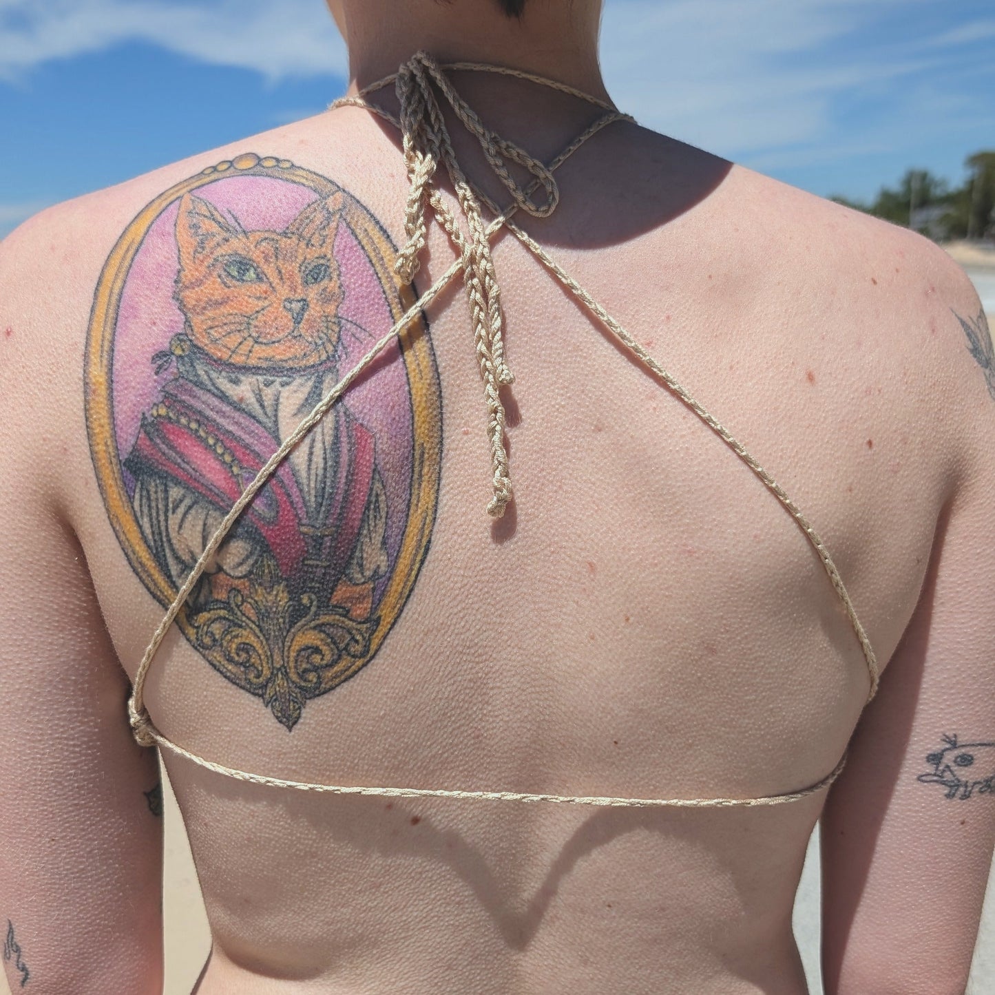 Person with a cat tattoo on their back at the beach, wearing cream bralette