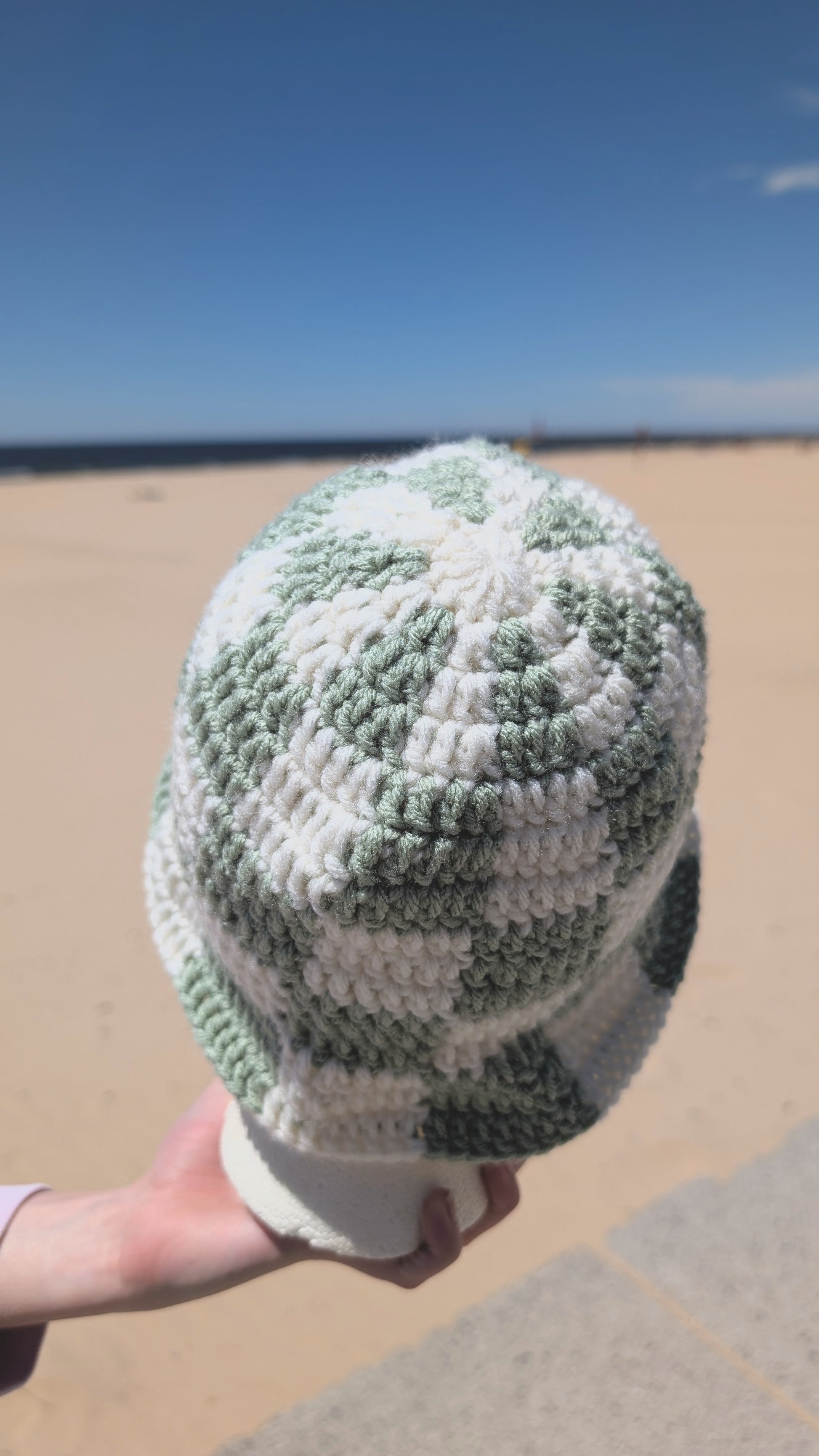 Green and white patterned beanie held by a hand on a beach.