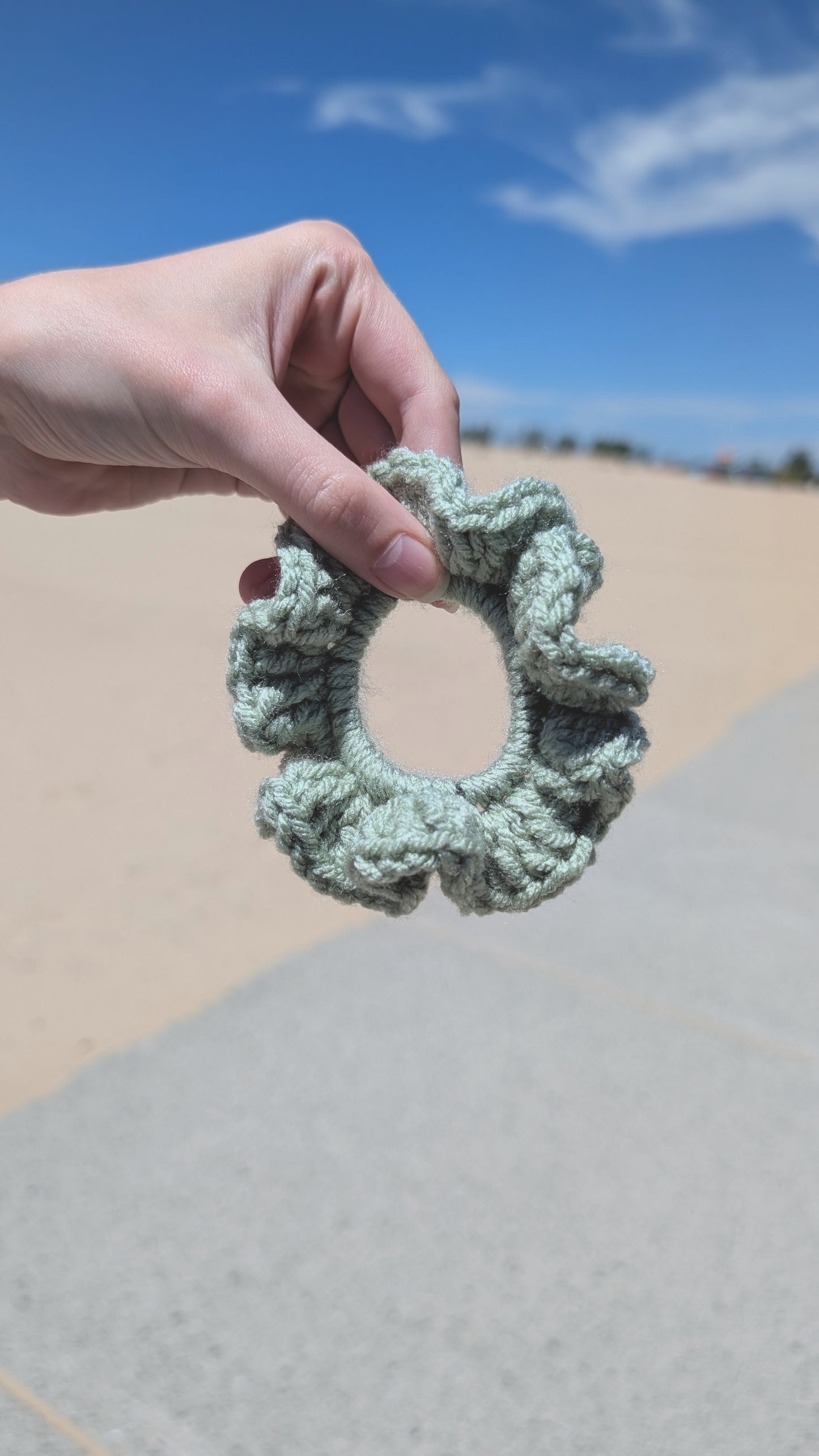 Hand holding a green crochet scrunchie against a blue sky and sandy background