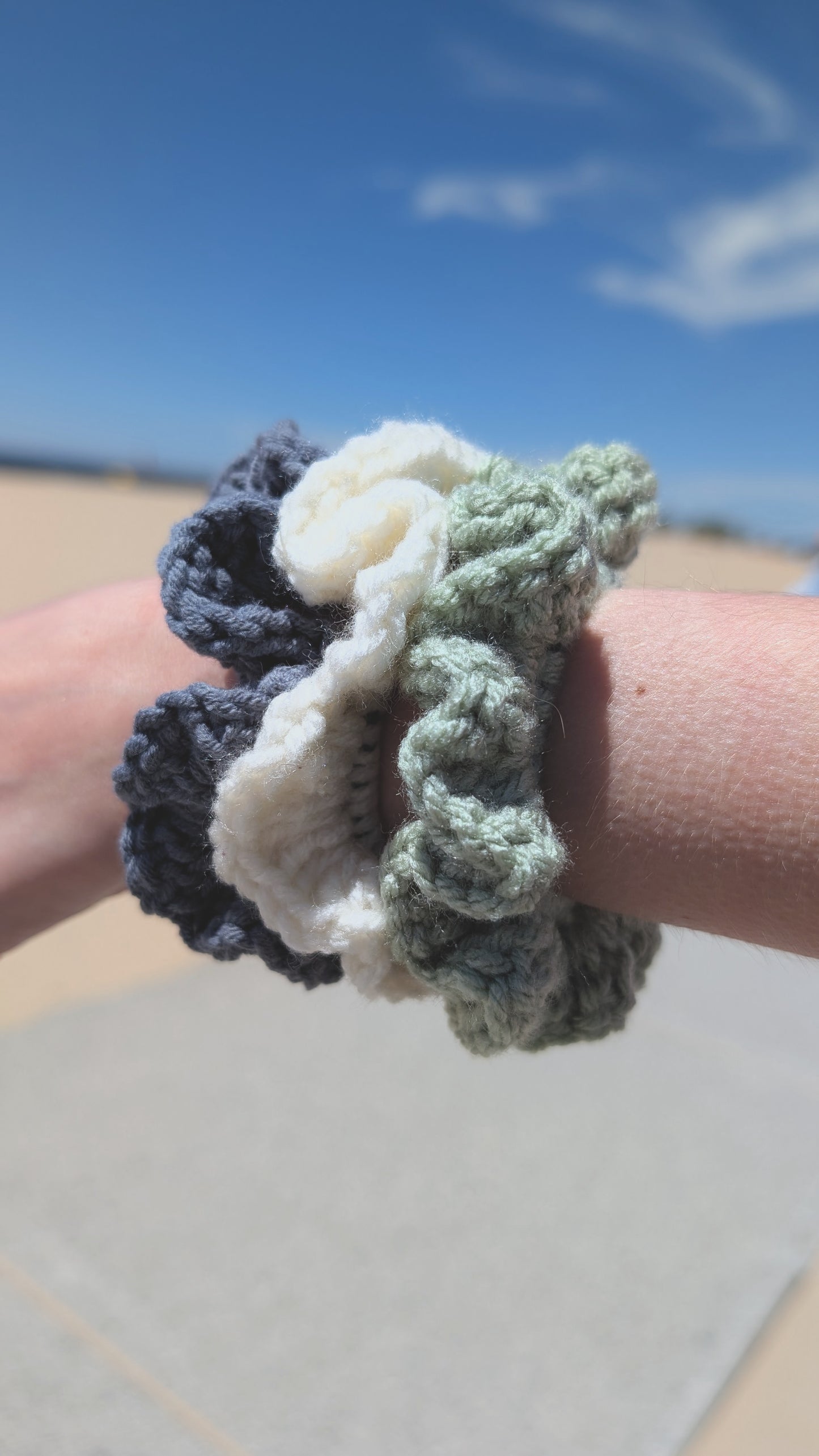 Three crocheted scrunchies on a wrist with a beach in the background